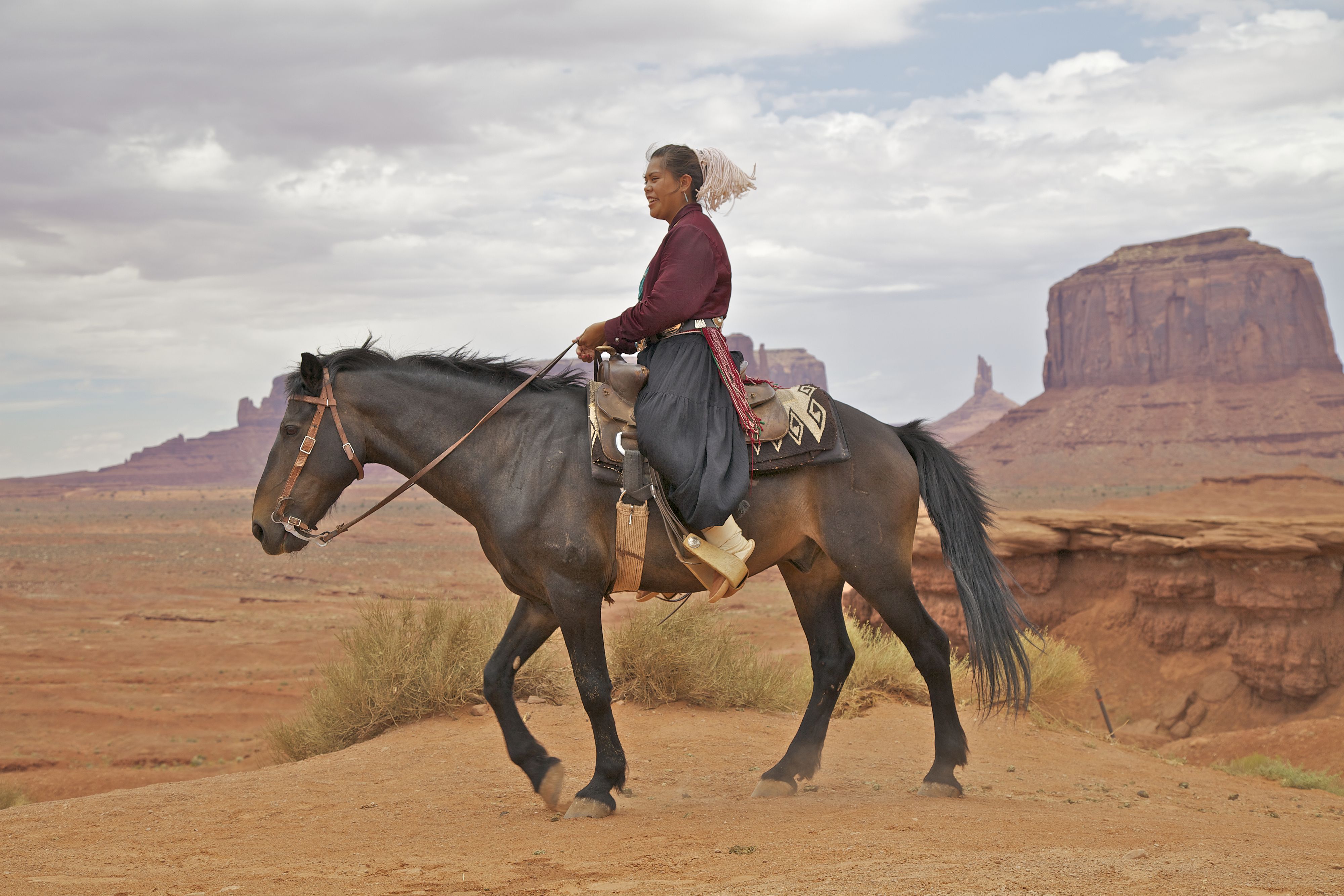 Person riding a dark horse across a reddish desert, with large sandstone mesas in the background and the rider wearing a maroon jacket and scarf.