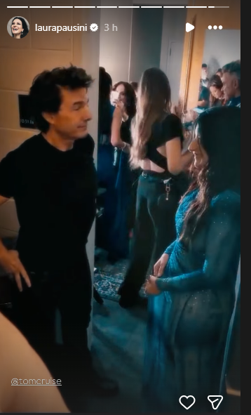 Instagram Story frame: a man in black chats with a woman in a shimmering blue dress in a backstage hallway, with other guests in the background.
