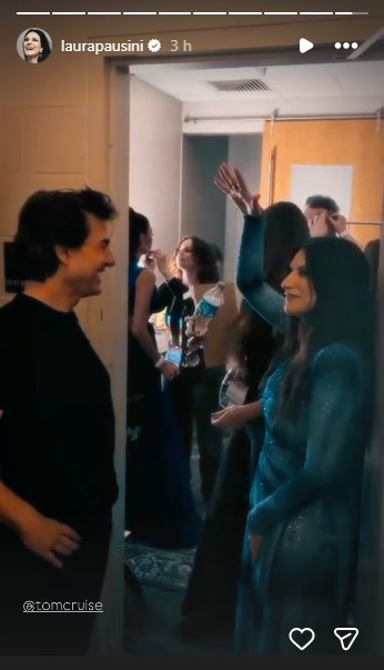 Backstage hallway greeting: a man in black chats with a group of people, a woman in a blue dress waves hello nearby.