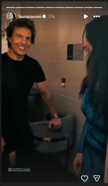 Two people smiling and chatting in a backstage hallway; man in a black T-shirt beside a woman in a blue dress by a sink area.