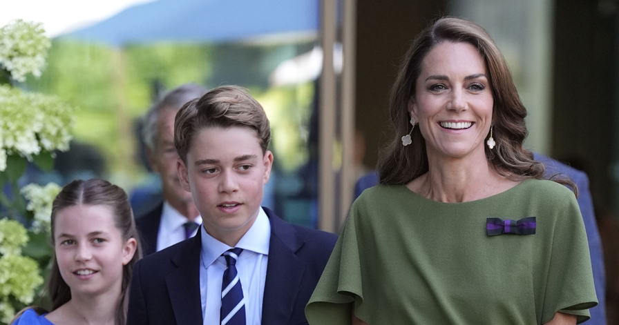 Smiling woman in a green dress with a bow walks beside a boy in a suit and a girl, exiting a building outdoors.