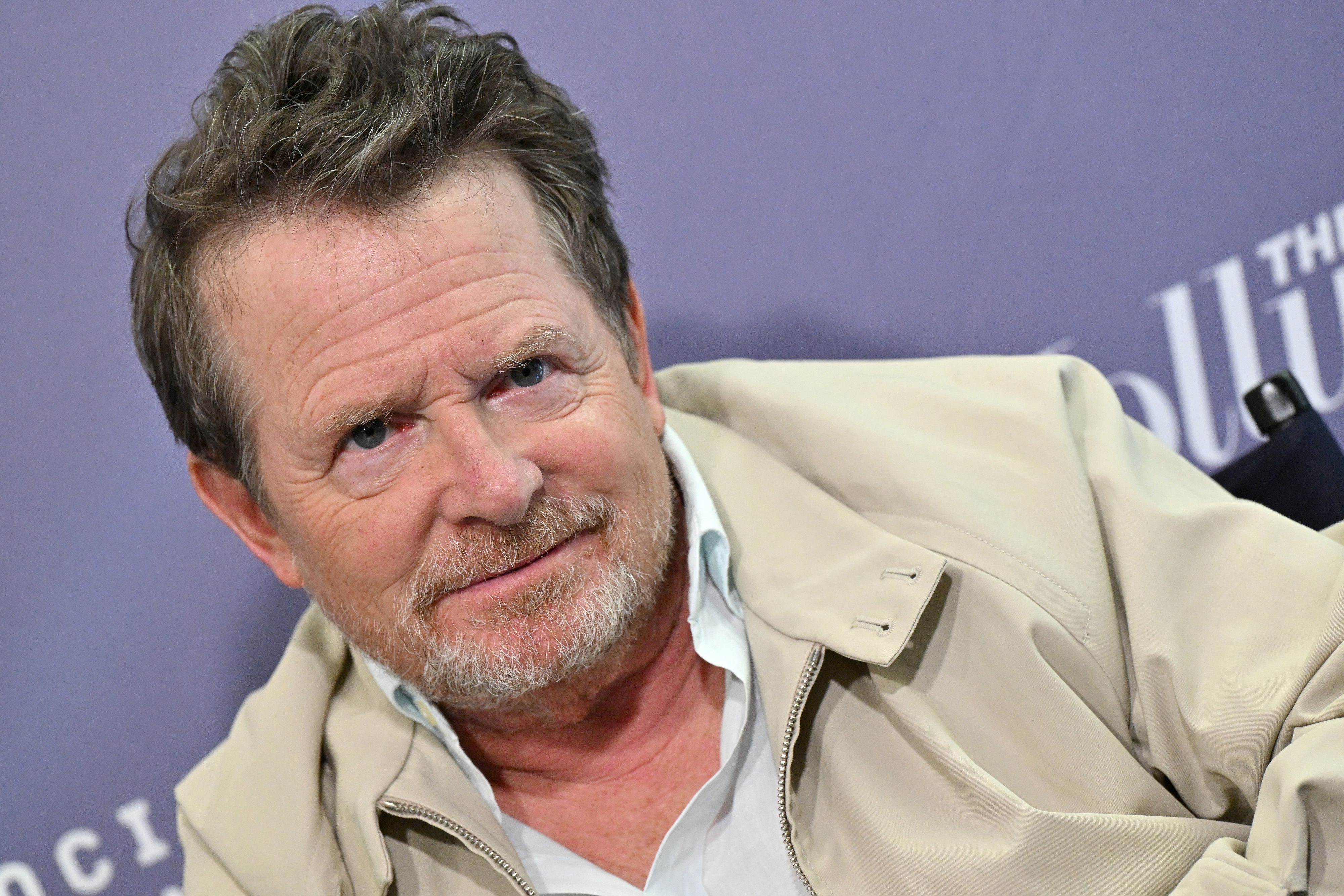 Close-up of a middle-aged man with a gray beard and mustache, wearing a beige jacket, tilted head against a purple backdrop.