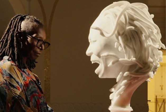 Woman with glasses in a colorful jacket studies a large white sculpture bust with flowing, rope-like hair in a gallery.