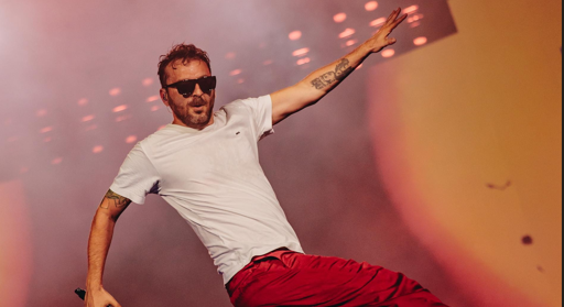 Male performer in a white T-shirt and red shorts dancing on stage, wearing sunglasses with tattooed arms under colorful stage lights.