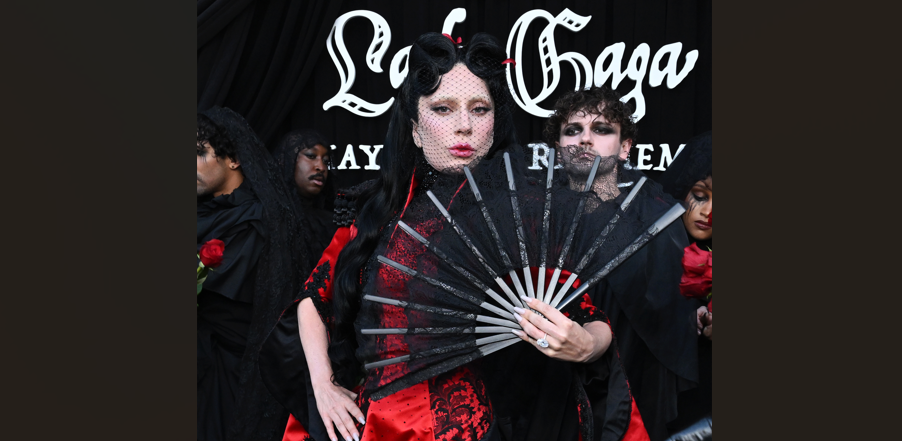Performer in a red and black lace dress, holding a black folding fan with a veil, in front of a Lady Gaga backdrop.