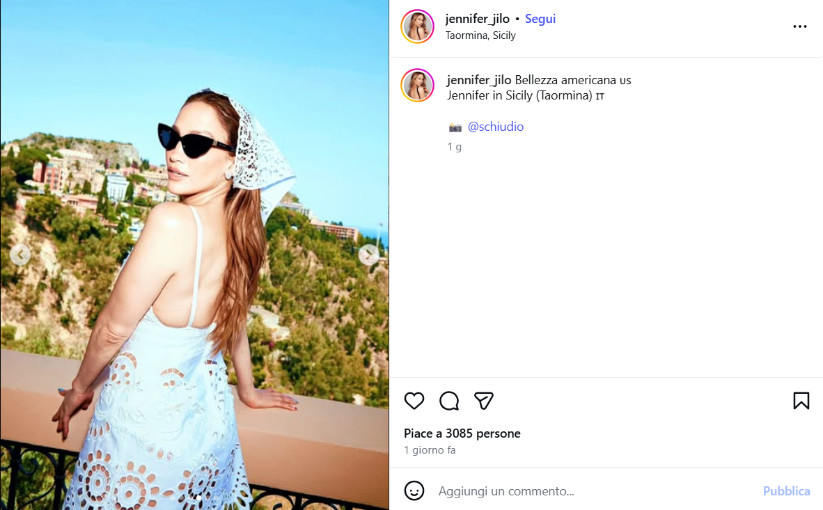 Woman wearing a white lace dress and black sunglasses posing on a balcony overlooking a hillside town under a clear blue sky in Sicily, Taormina perspective.