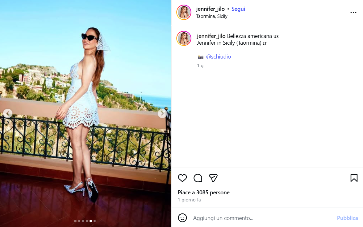 Woman in a light-blue lace dress and sunglasses stands on a balcony with a coastal Sicilian town and blue sky behind.