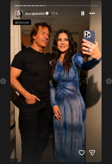 Two people pose for a backstage selfie: man in a black t-shirt and a woman in a blue dress, a phone visible in the foreground.