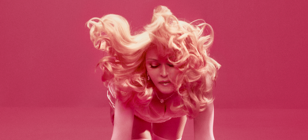 Portrait of a blonde woman bending forward with voluminous curls flowing around her, pink lighting tinting the scene.