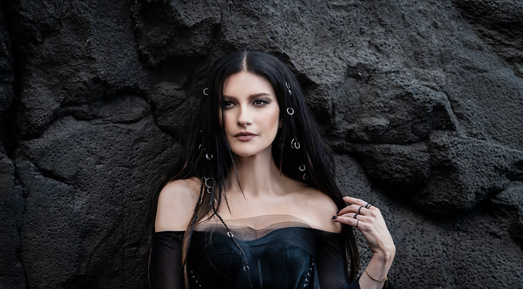 Portrait of a woman with long black hair wearing an off-shoulder black top against a dark rocky wall