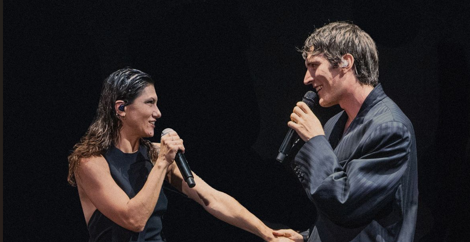Two performers on stage share a microphone moment, facing each other and clasping hands while singing.