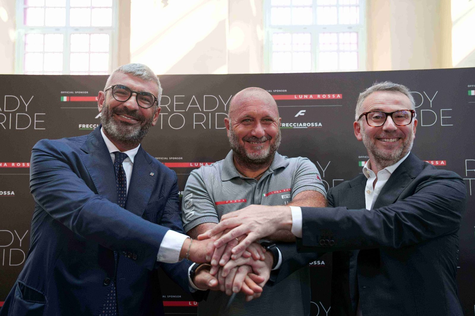Three men in suits join hands in a triple-hands stack at a business event, smiling for the camera in front of a sponsor backdrop.