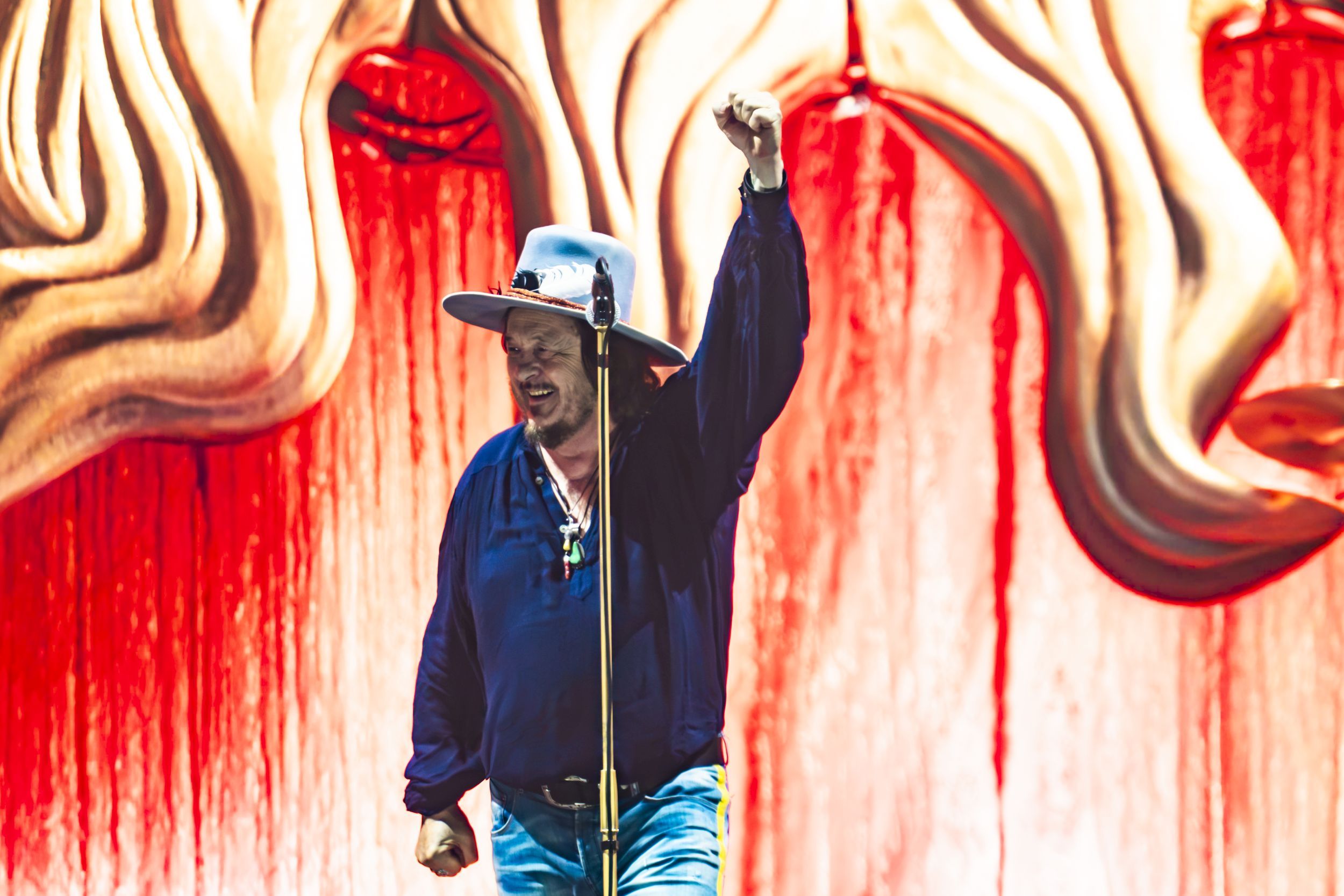 Man on stage wearing a light blue cowboy hat and dark shirt raises his clenched fist as he performs, with bright red curtains in the background.