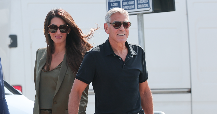 Smiling man in a black polo with sunglasses walking beside a woman in a green blazer and sunglasses.