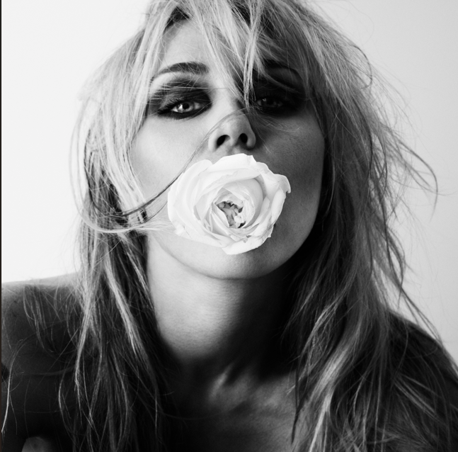 Black-and-white portrait of a woman with a white rose clenched in her mouth, tousled hair and smoky eyes.