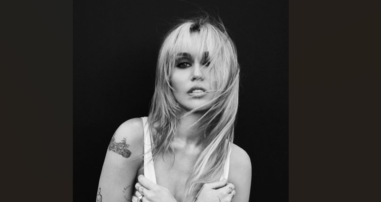 Black-and-white portrait of a woman with long blonde hair, wearing a white tank top, tattoos visible on her arm.