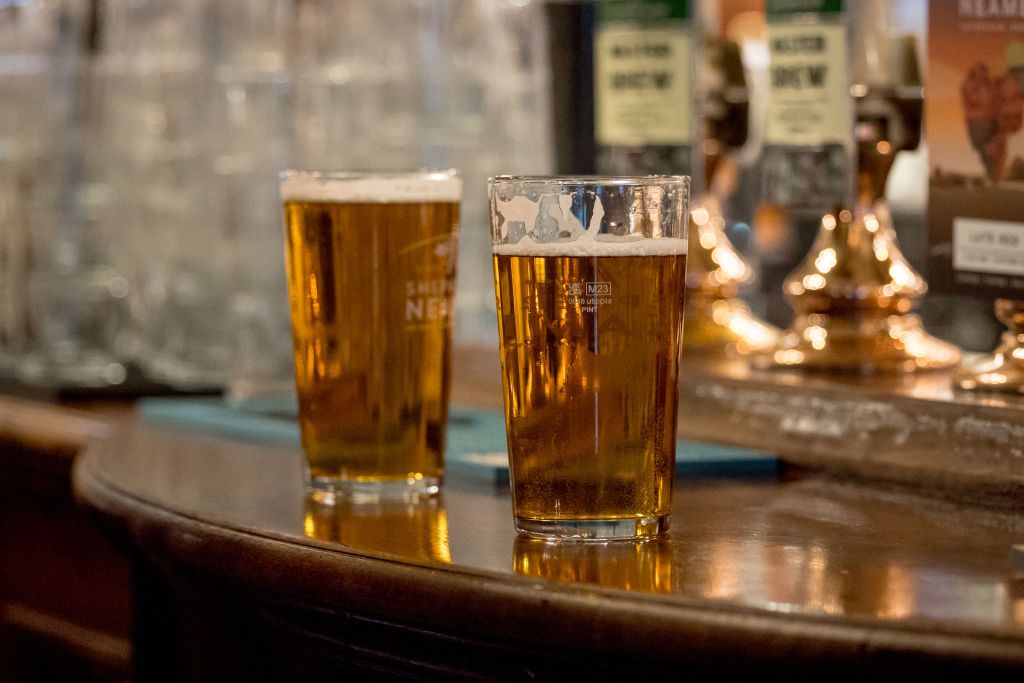 Two pint glasses of beer on a wooden bar counter with foamy tops and reflections in the surface, background taps visible.