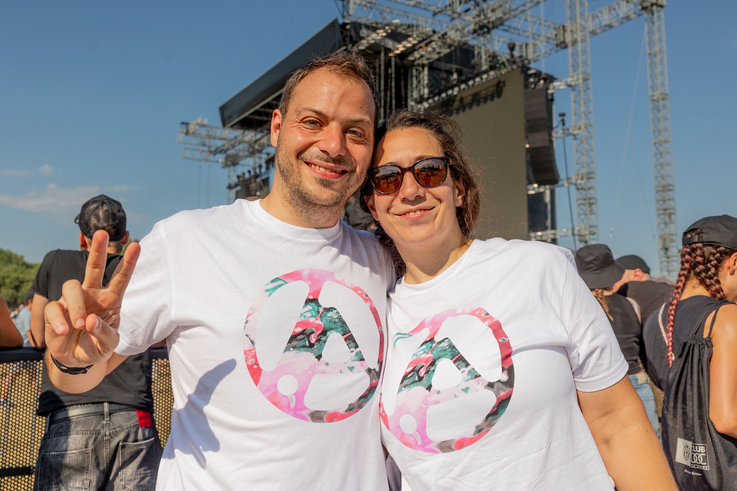 Smiling man and woman pose for a selfie at an outdoor concert, with a large stage behind them and blue sky.