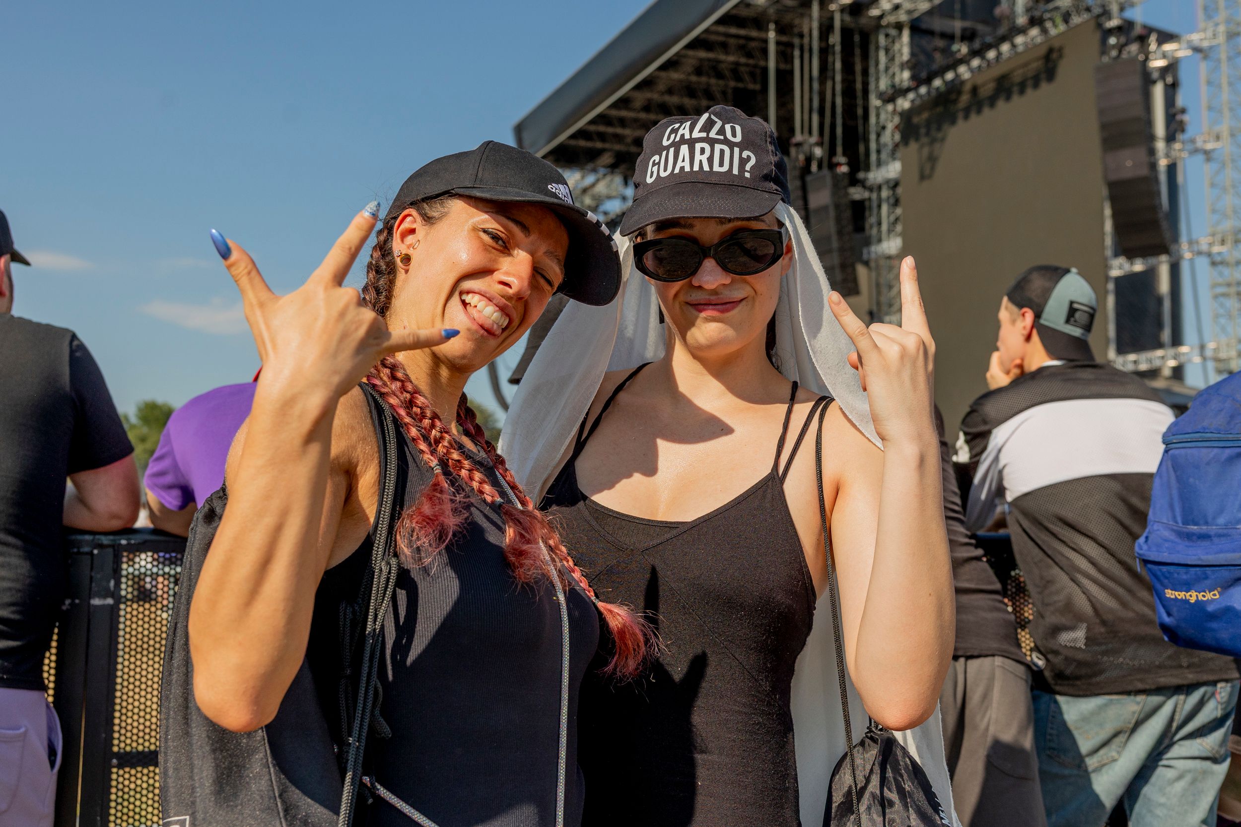 Two friends at an outdoor concert, smiling and making the rock hand sign with a large stage behind them.