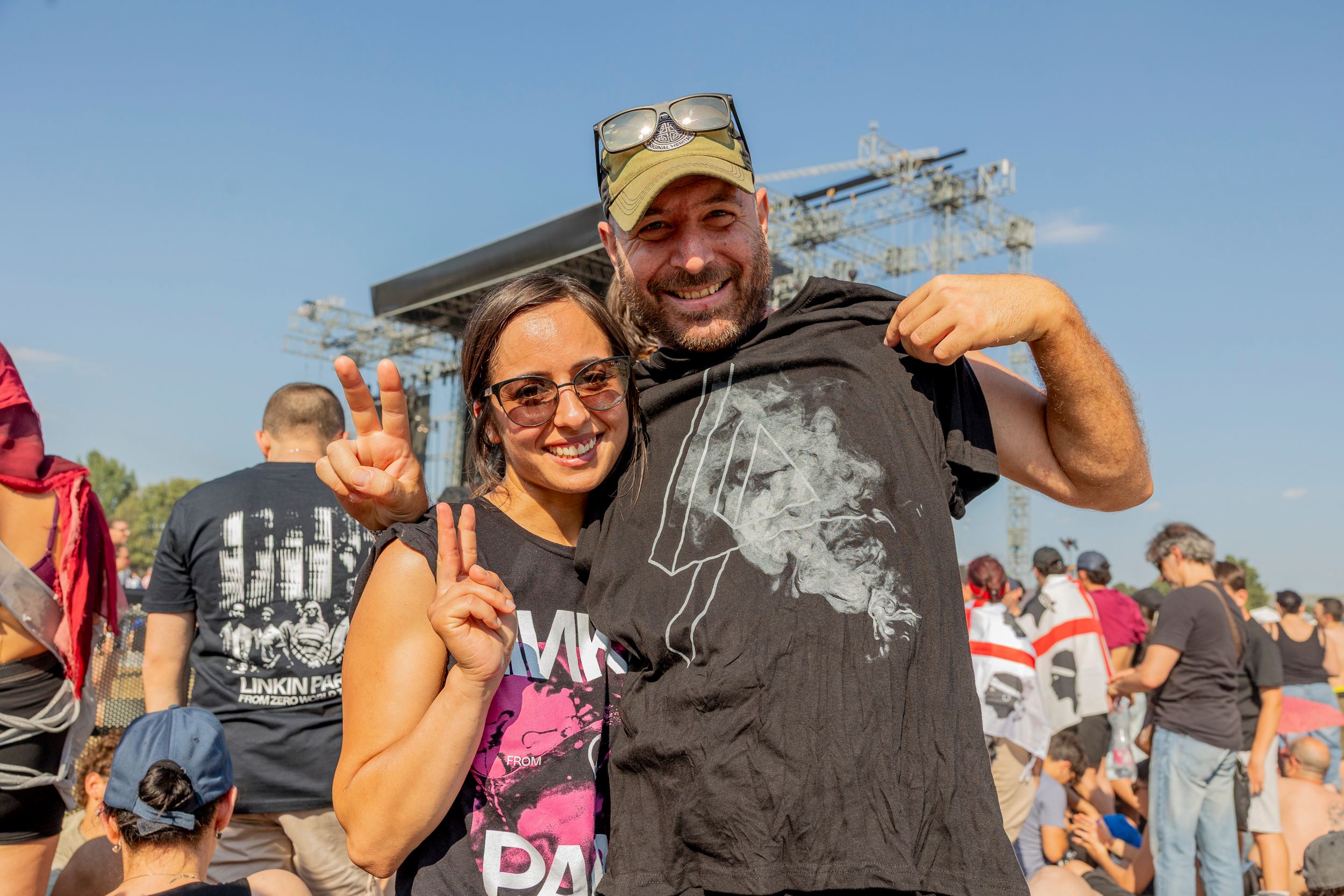 A smiling man and woman pose together, making peace signs at a sunny outdoor concert with a large stage and cheering crowd behind them.
