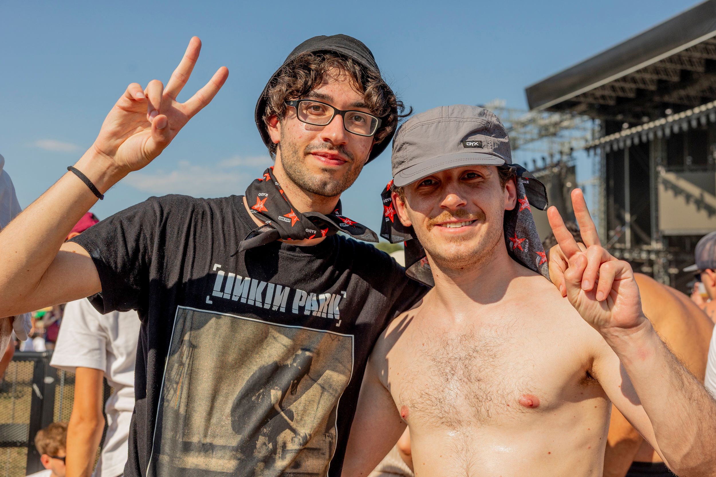 Two men posing at an outdoor festival, flashing peace signs; one in a black hat and glasses, the other shirtless with a gray cap, stage behind them.