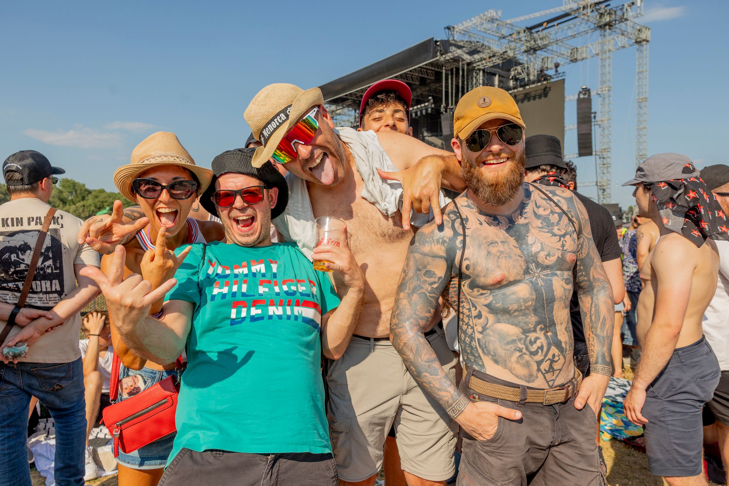 A group of smiling festival-goers posing for a photo in front of a large stage with rigging and lights. People wear hats, sunglasses, and colorful clothing under a clear sky.
