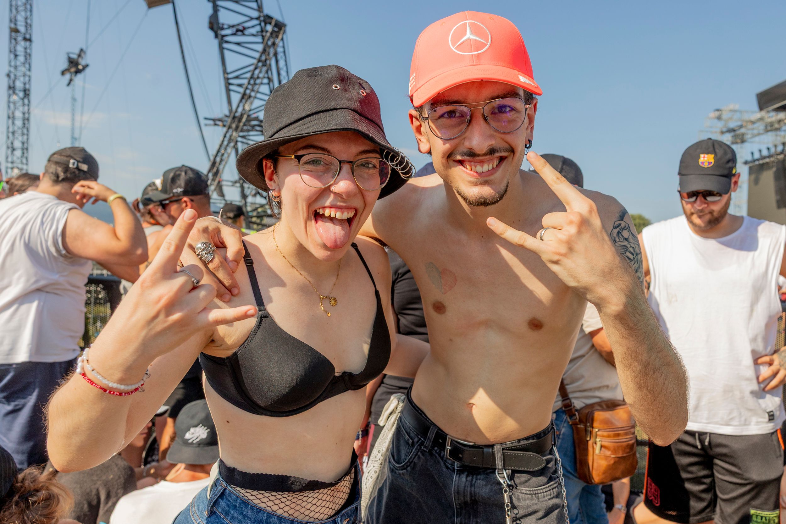Two young friends at an outdoor festival pose for a photo, wearing hats and sunglasses with a crowd and stage rig in the background.