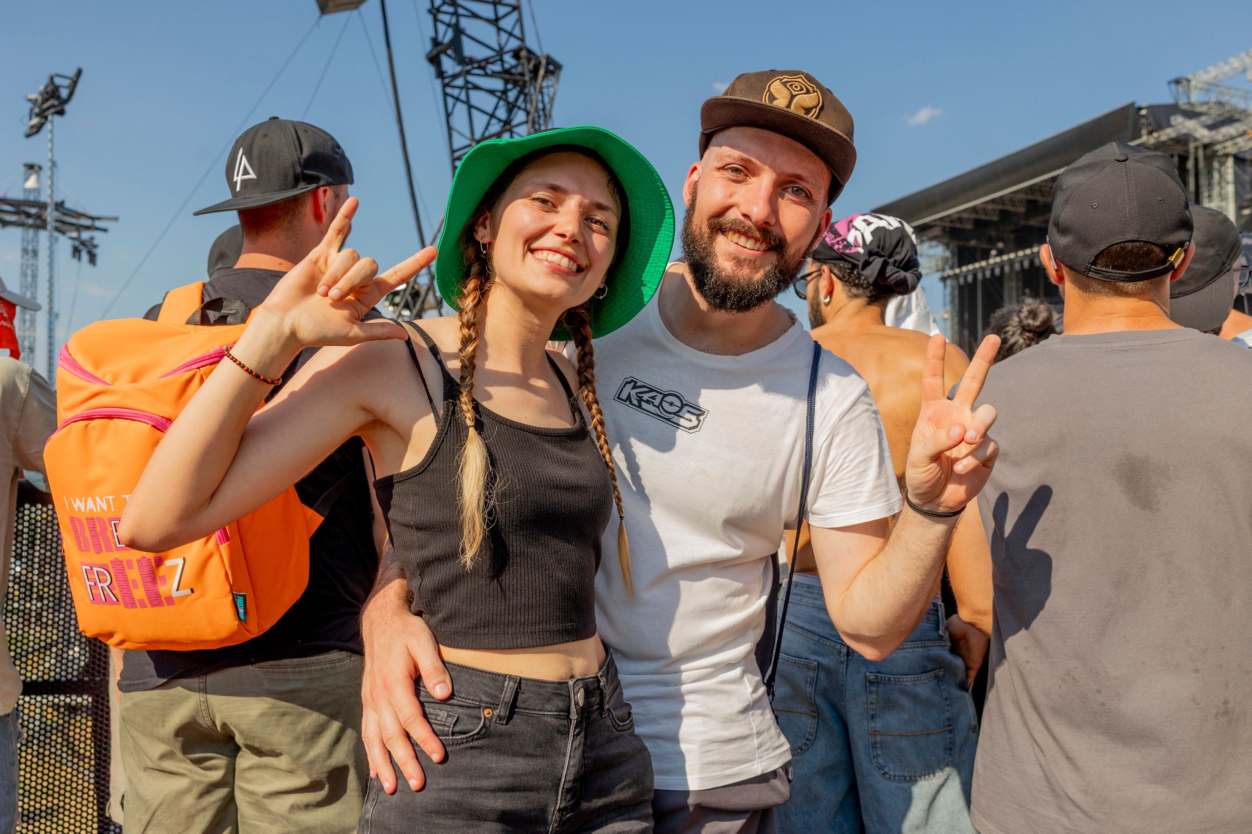 Group of friends at an outdoor concert posing for a photo, smiles, and rock hand gestures, with stage equipment in the background.