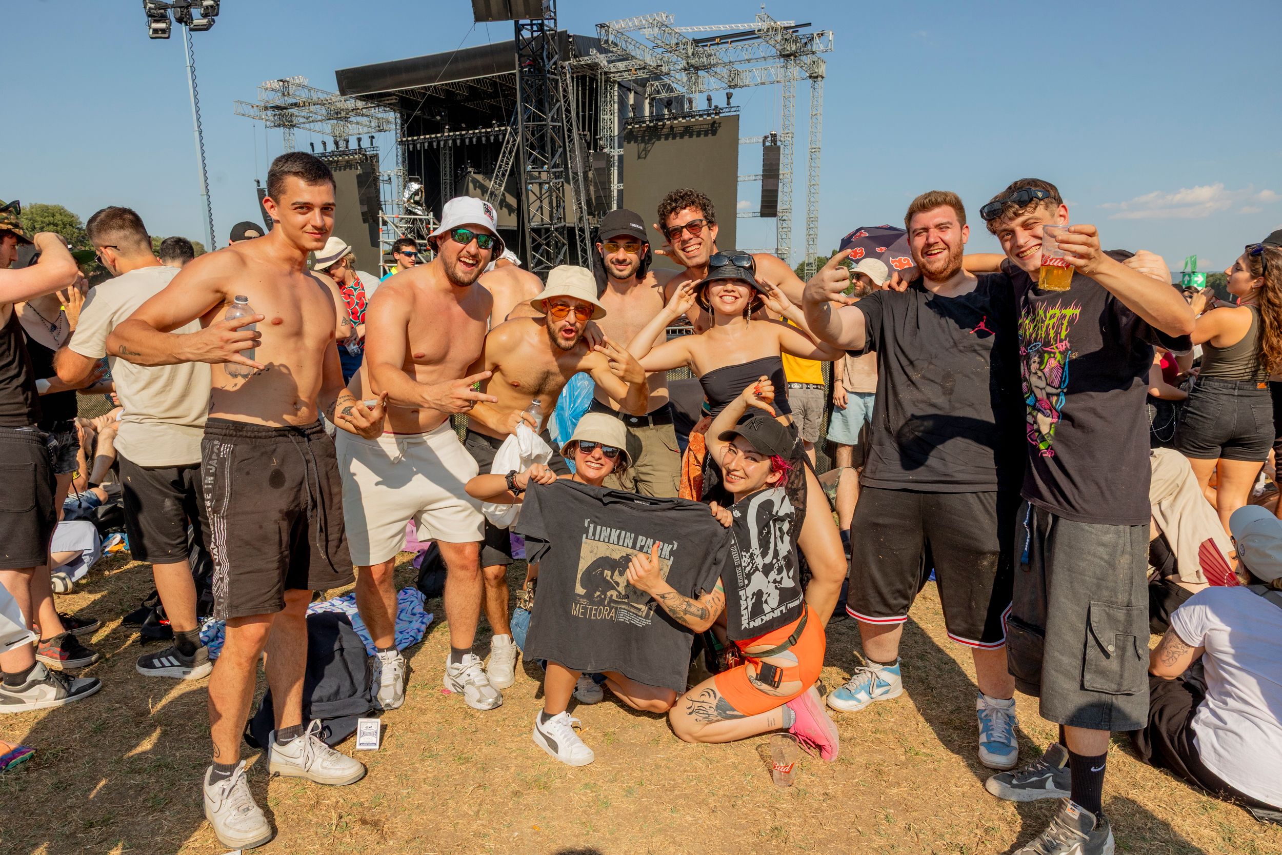 Group of friends posing for a photo at an outdoor music festival with a large stage behind them behind scaffolding and lights.