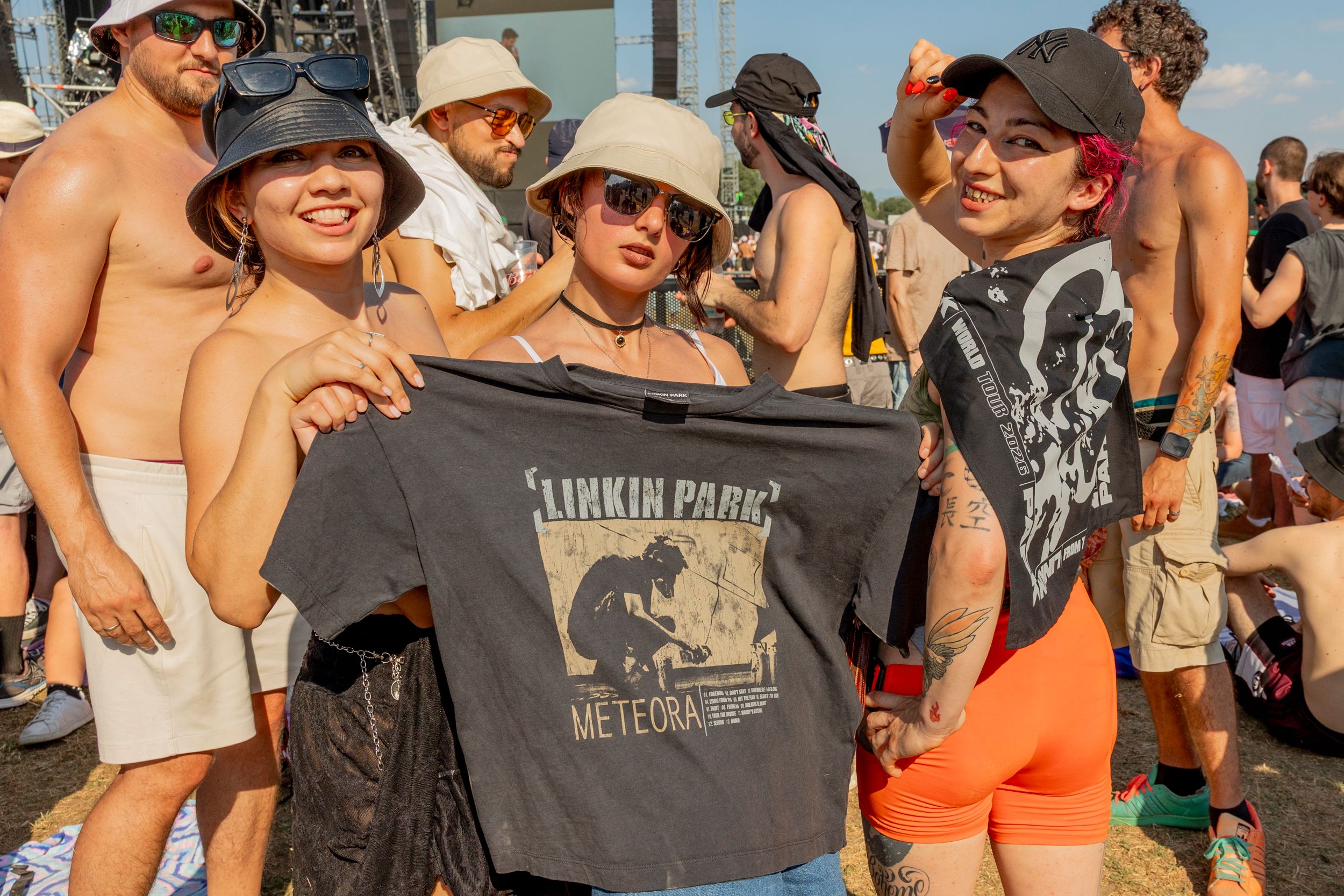 Smiling group of festivalgoers posing for a photo, several wearing hats and sunglasses, holding a Linkin Park T-shirt.