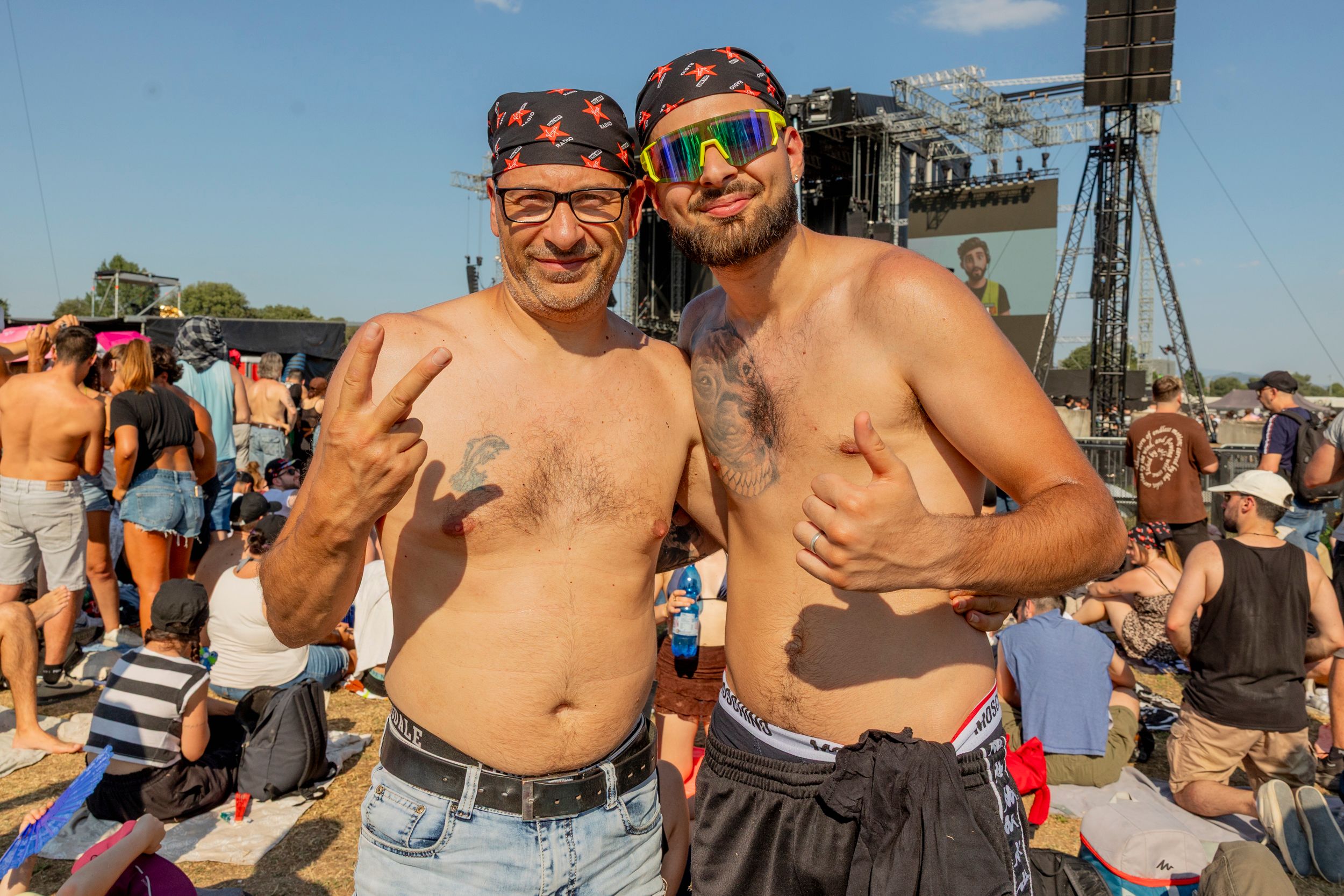 Two shirtless men posing with their arms around each other at an outdoor festival, crowd and stage behind them.