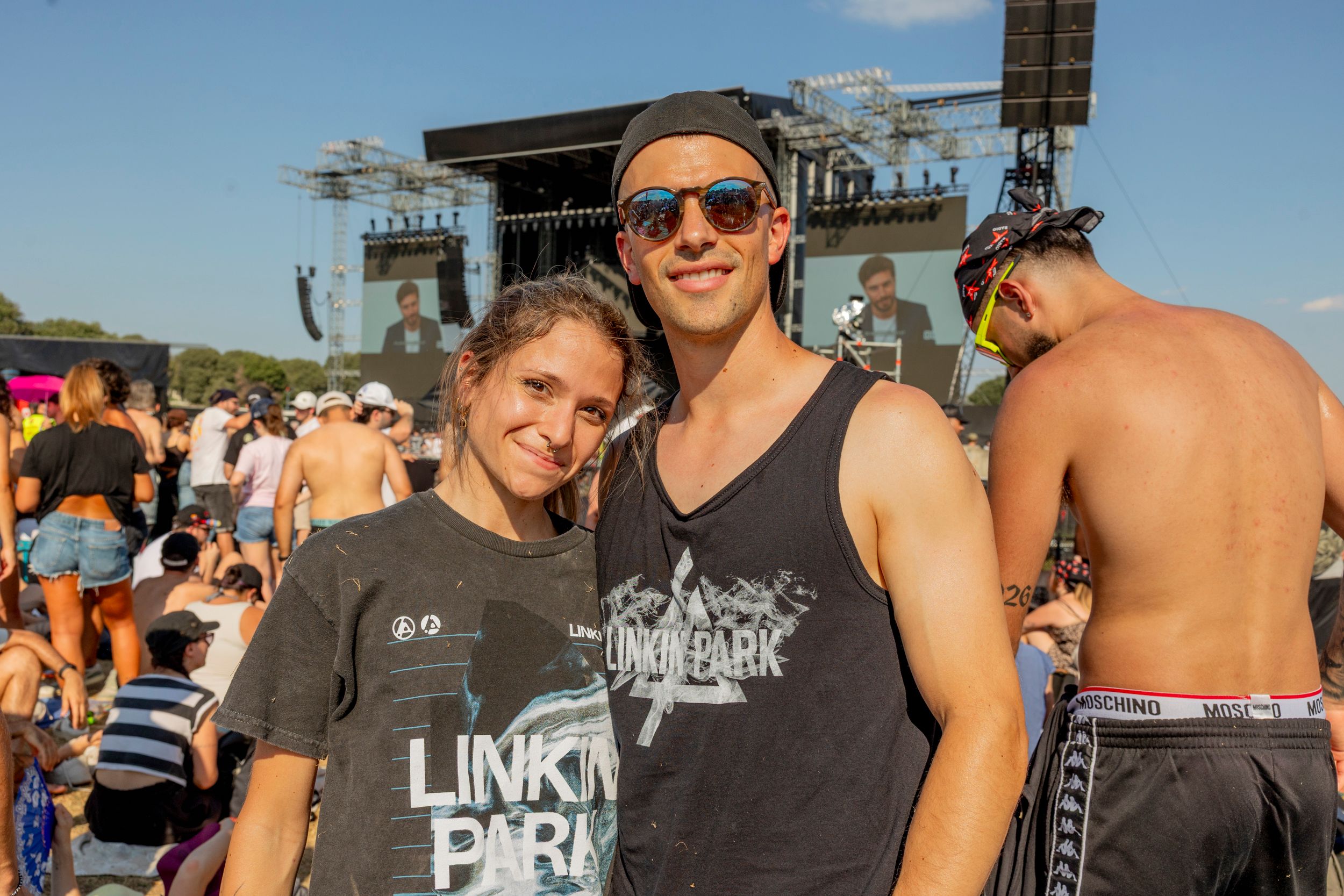 Two young people pose for a photo at an outdoor concert with a large stage and cheering crowd behind them.