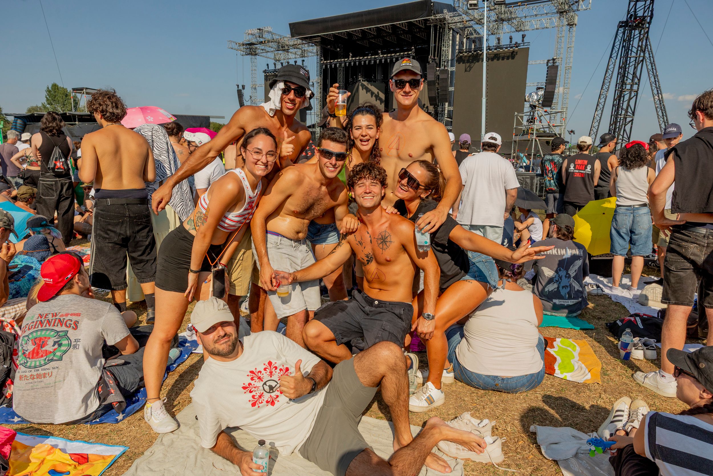 Group of friends posing for a photo in front of a large outdoor festival stage, sunny day, drinks in hand.