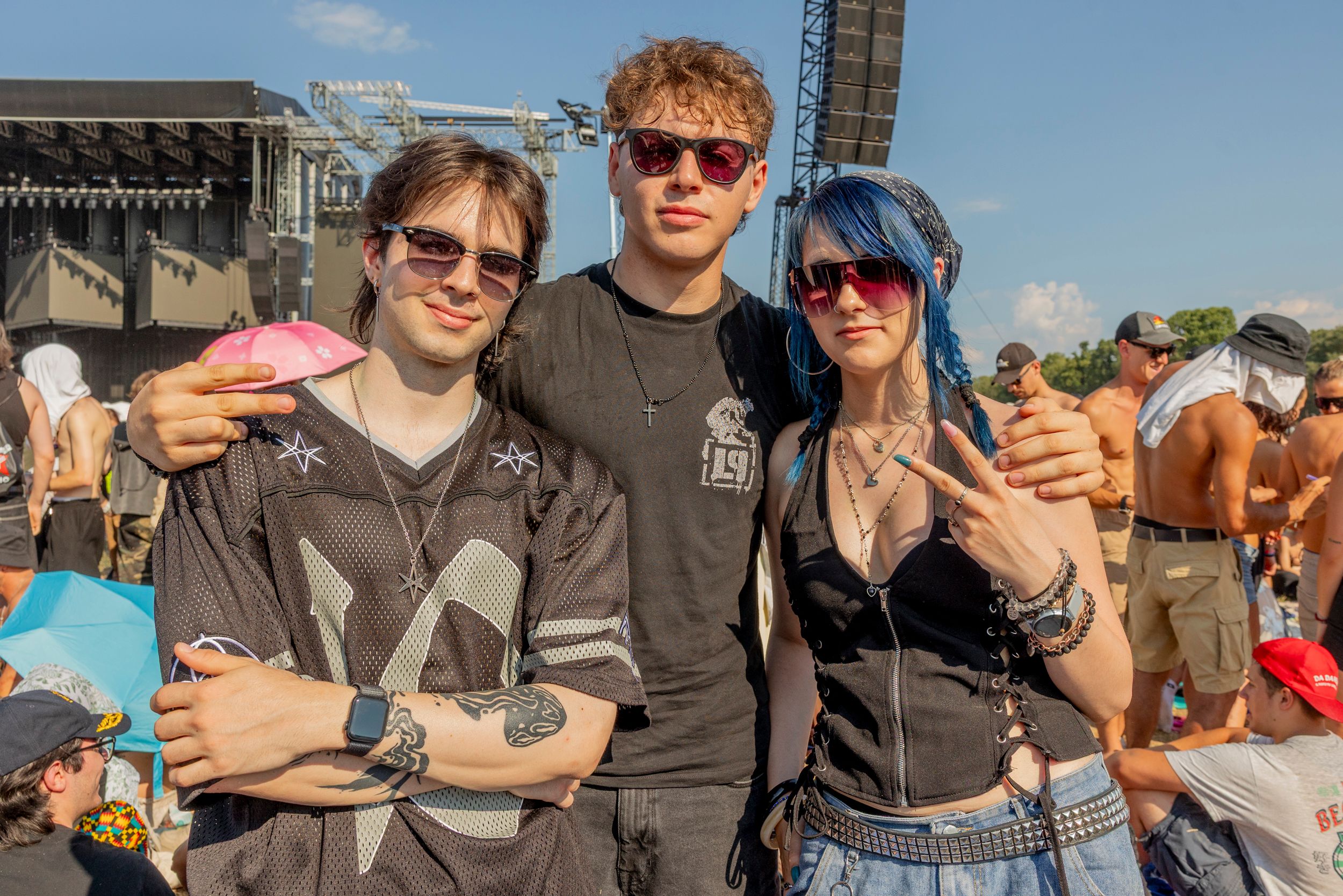 Three friends posing at an outdoor music festival, wearing sunglasses with a large stage behind them.