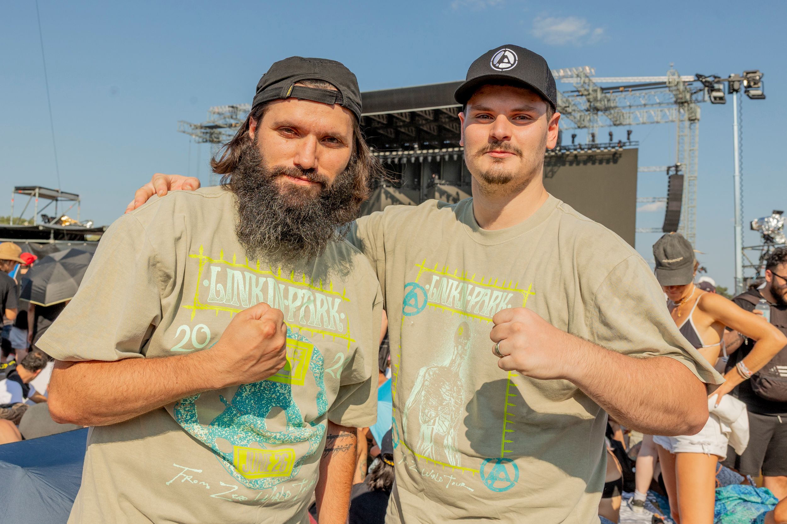 Two men pose for a photo with fists raised at an outdoor concert, wearing matching beige graphic T‑shirts. They stand in front of a large stage rigging.