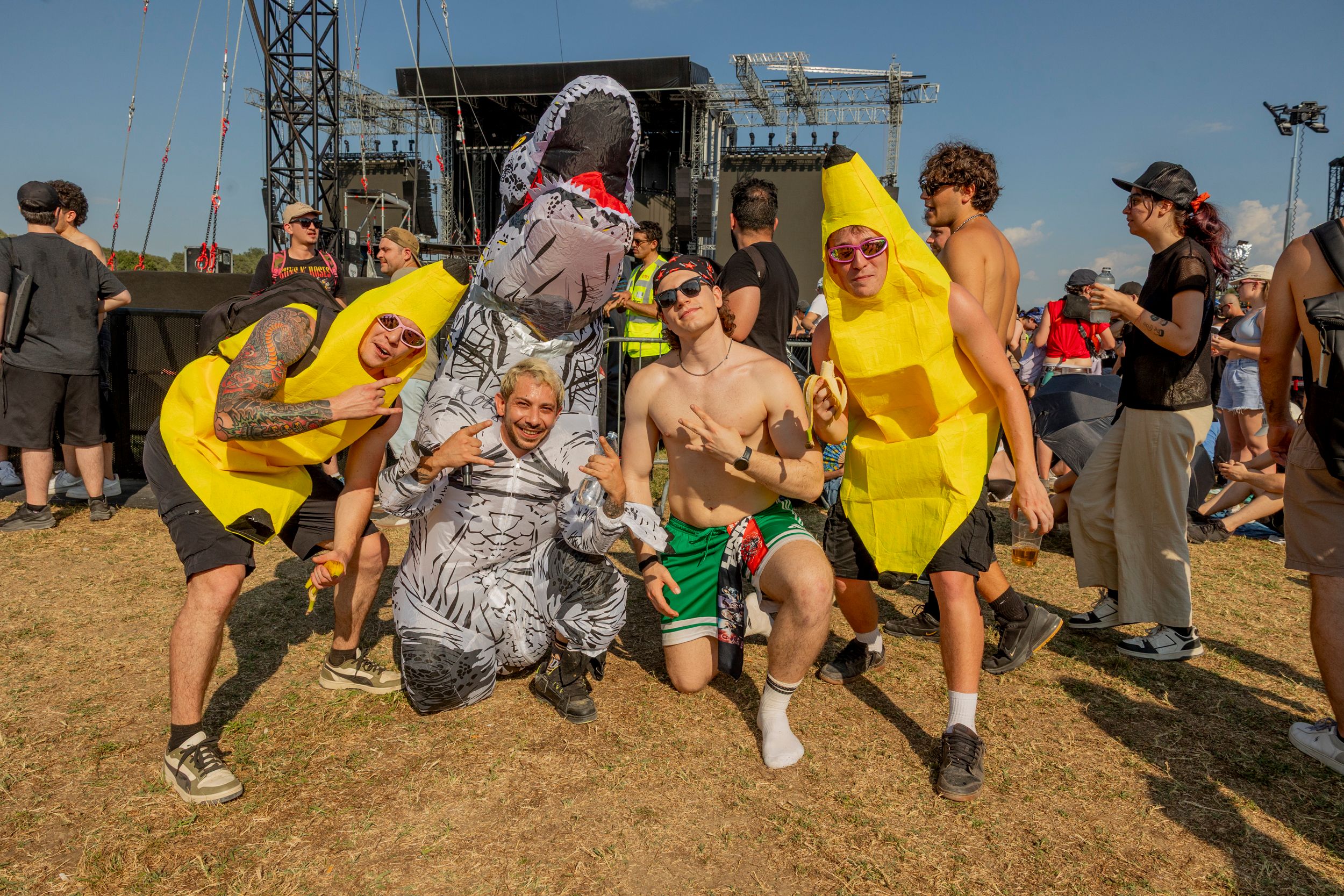 Group of festival-goers posing in banana costumes and a dinosaur suit in front of a large stage on a sunny day at an outdoor music festival.