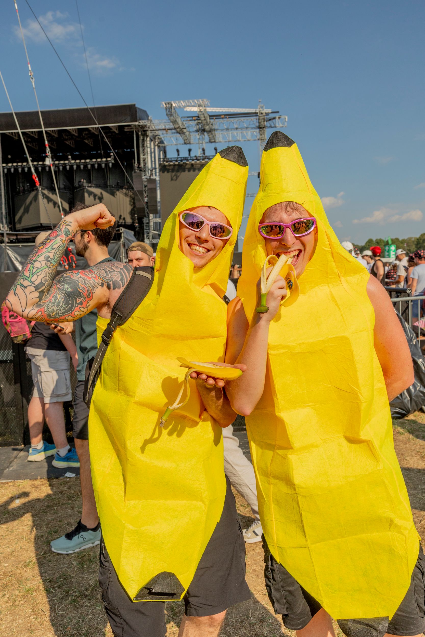 Two people in bright yellow banana costumes and pink sunglasses posing at an outdoor concert with a stage behind them.