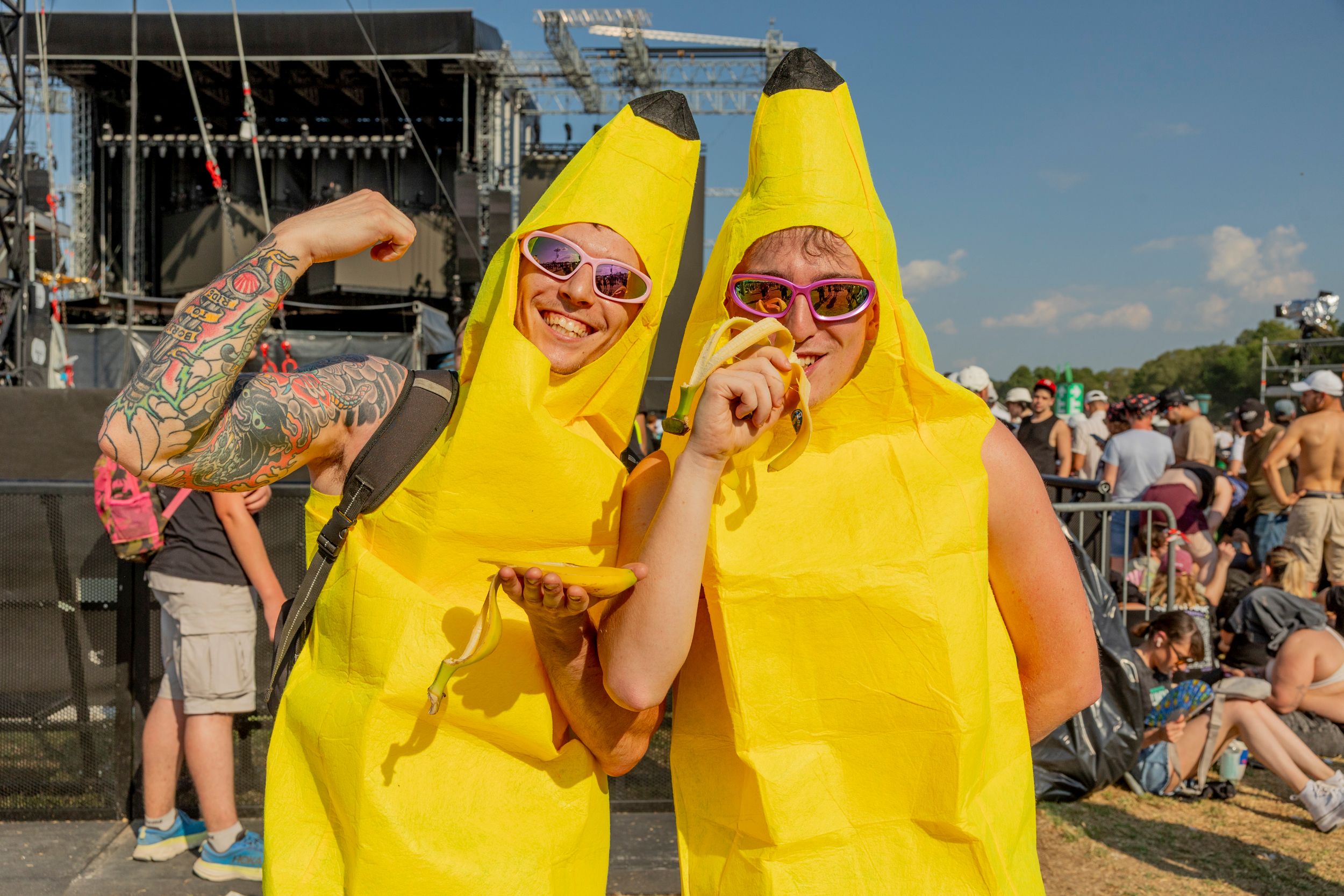 Two people in bright yellow banana costumes pose for the camera at an outdoor concert, wearing pink sunglasses and smiling.