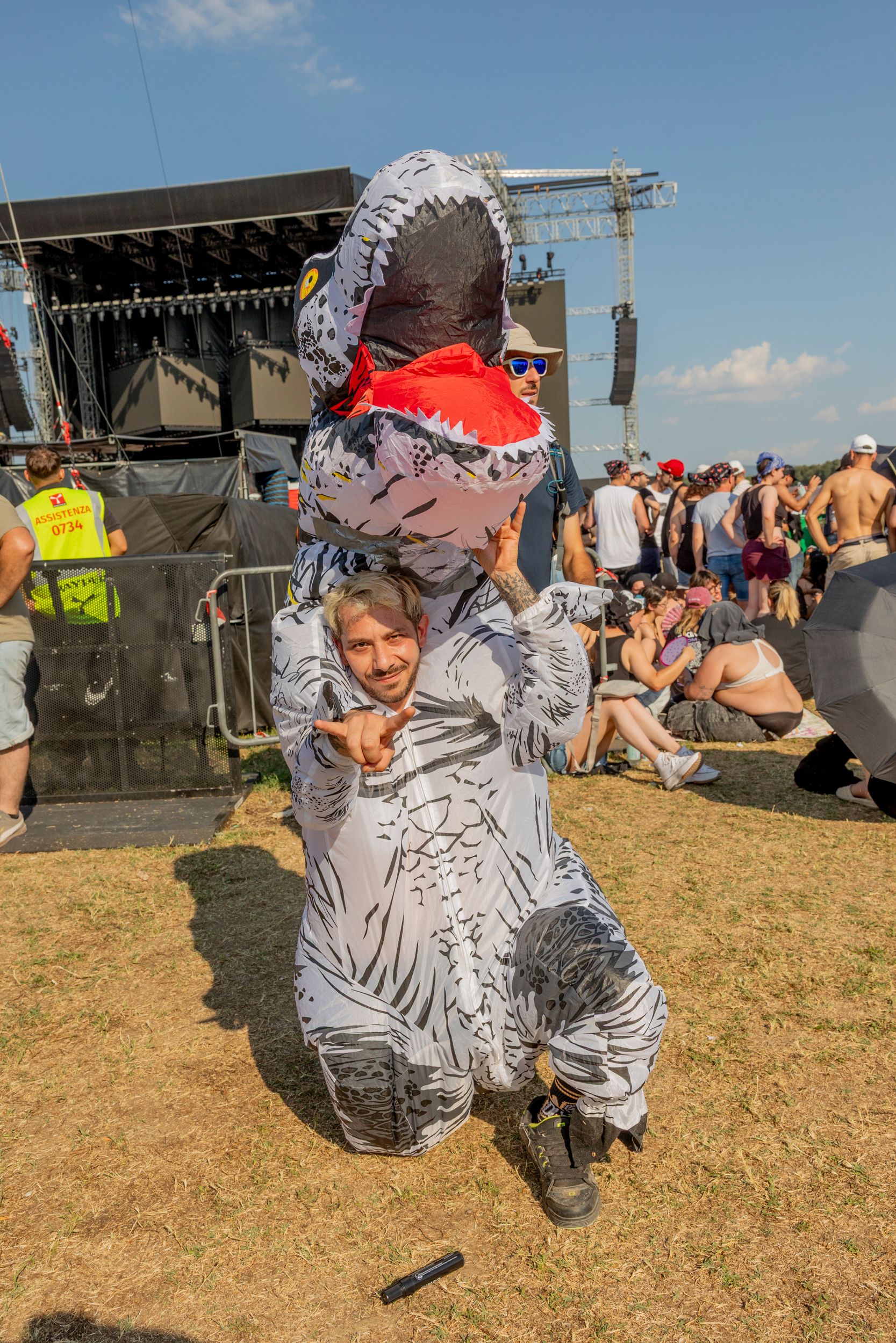 Person wearing a gray inflatable dinosaur costume poses on a grassy festival ground with a large stage and crowd in the background.