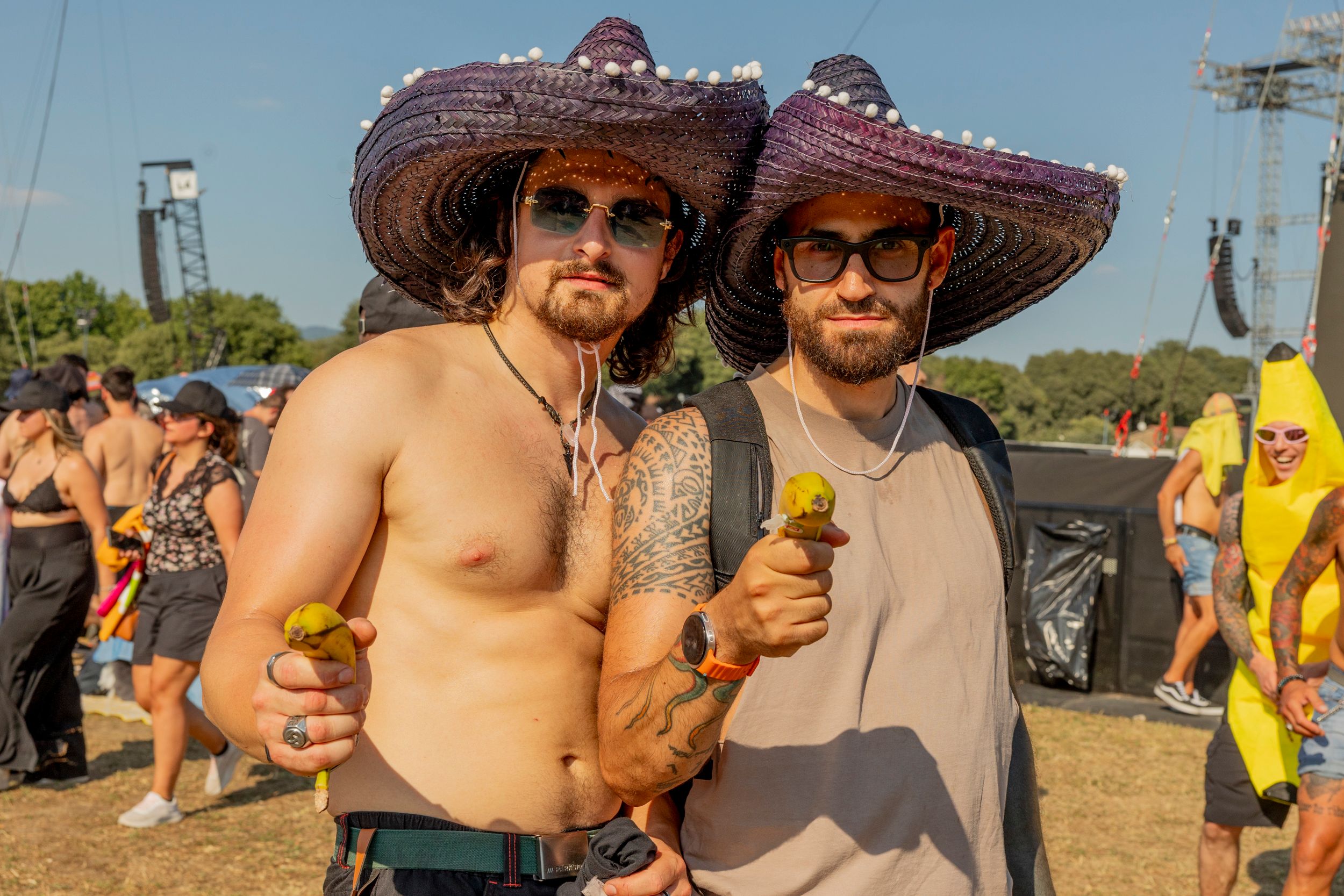 Two men with beards and tattoos wearing oversized purple sombreros and sunglasses pose for the camera at a sunny outdoor festival.