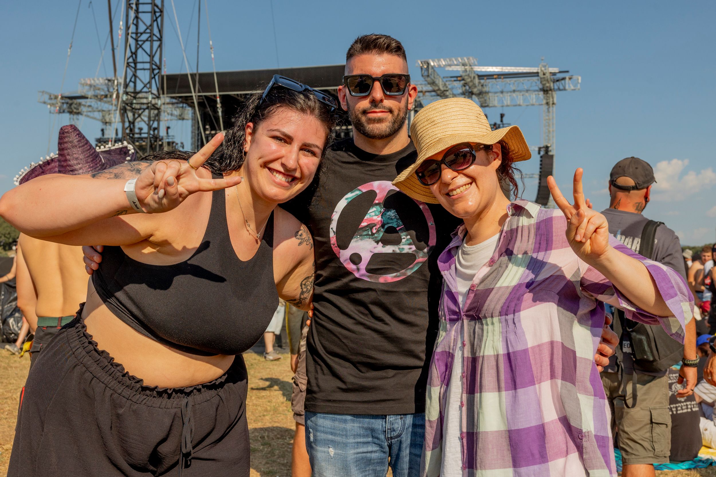 Three friends posing for a selfie at an outdoor concert, smiling and making peace signs with a festival stage behind them.