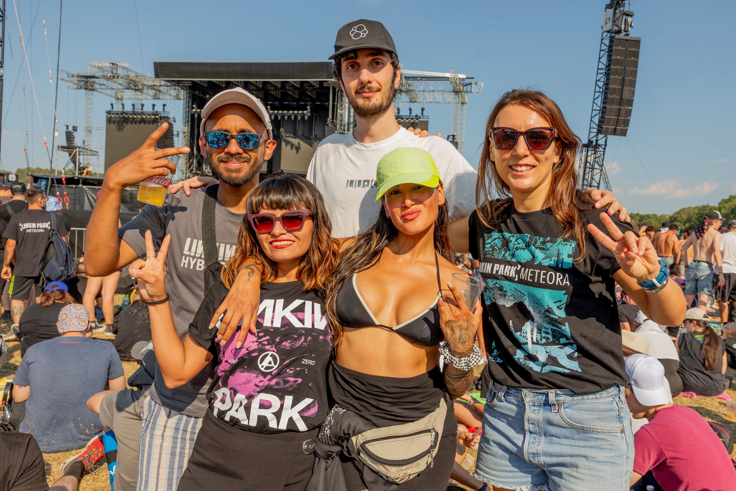 Group of five friends posing for a photo at an outdoor music festival, with a large stage behind them on a sunny day.