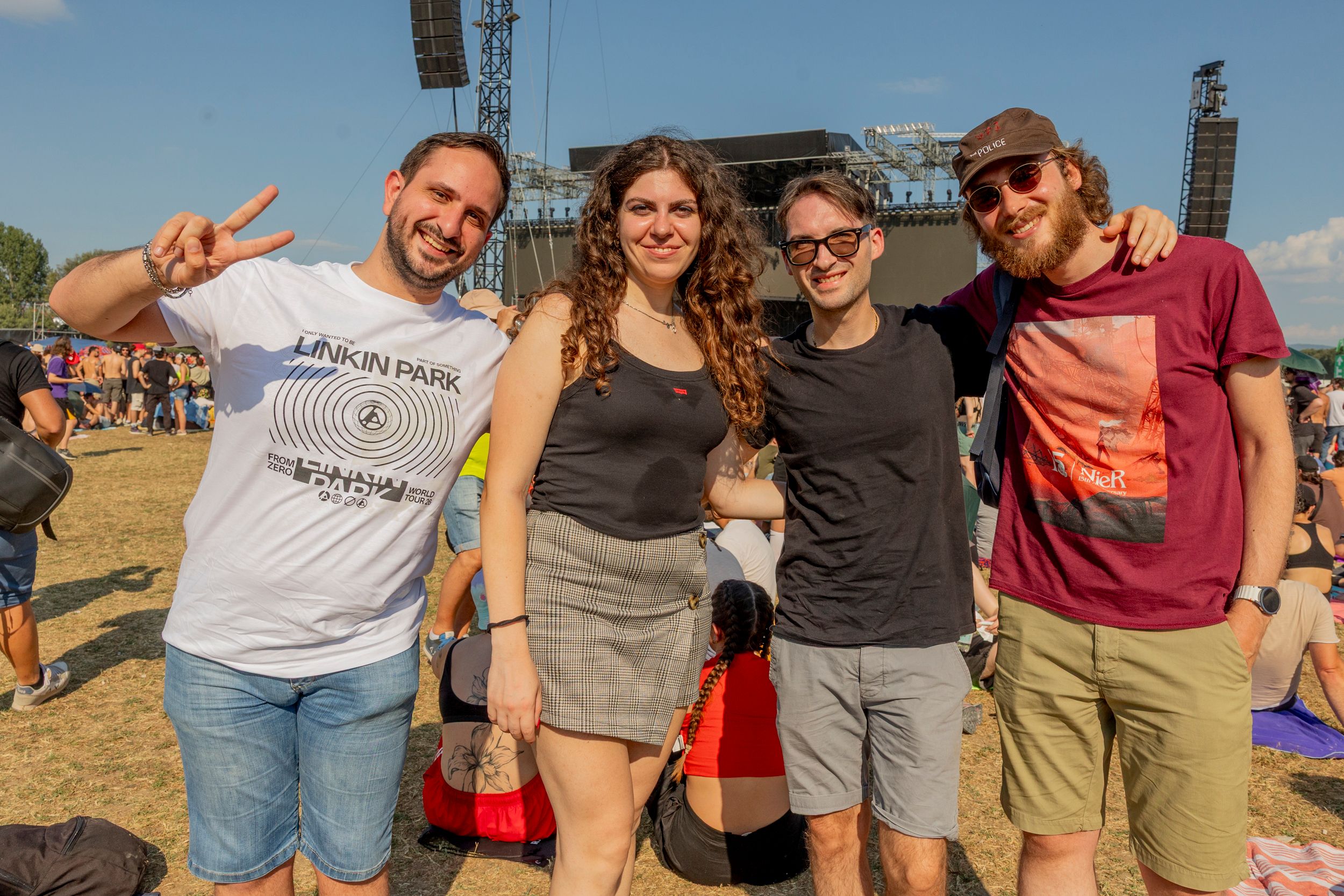 Four friends pose together for a photo at an outdoor music festival, with a large stage behind them on a sunny day.