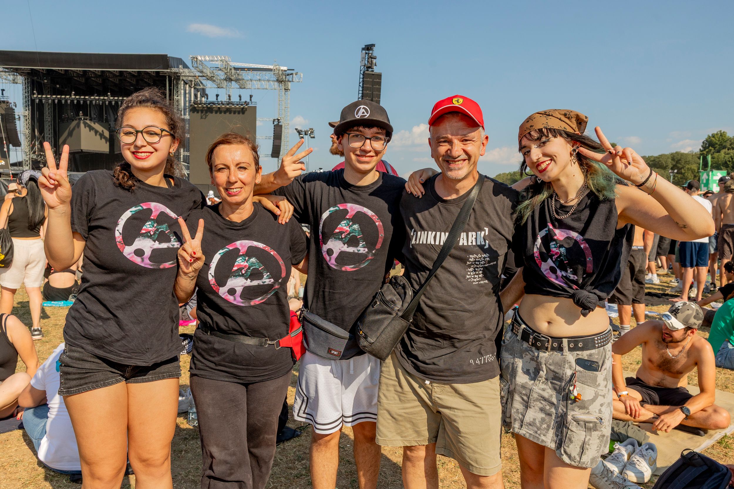 Five friends at an outdoor music festival pose together, wearing matching black shirts with a colorful circular logo and flashing peace signs in front of a stage.
