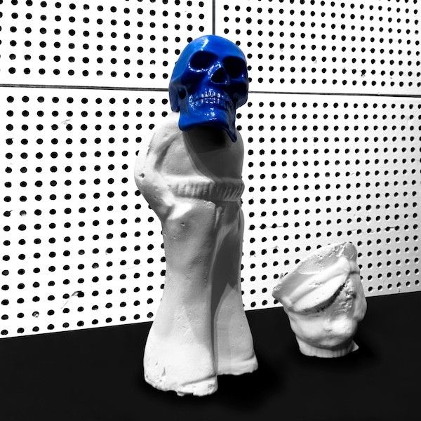 White ceramic hooded figure with a blue skull on its head stands on a black surface, a broken white cup nearby against a dotted wall behind them.