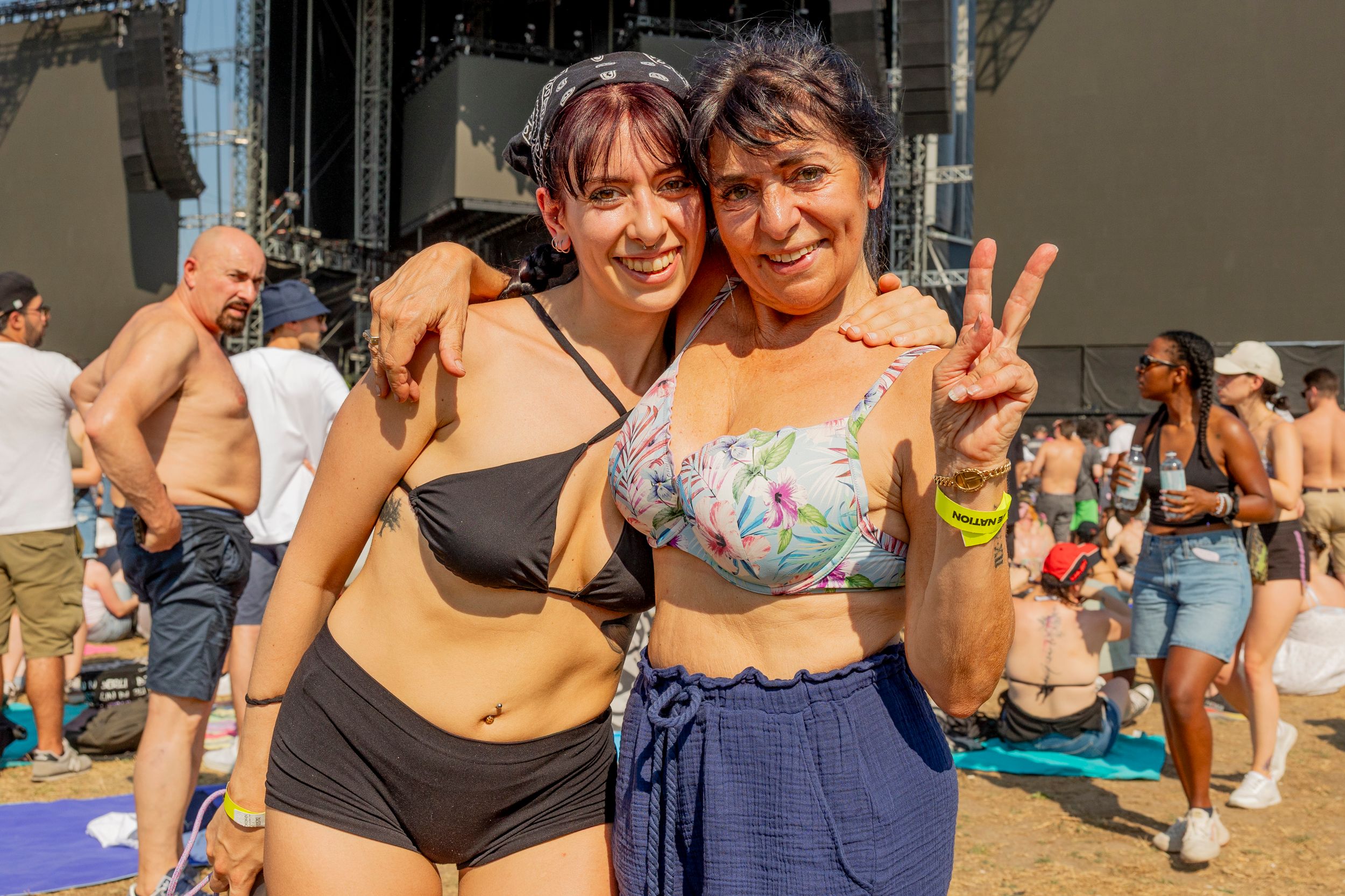 Two women in swimsuits pose together at a beach festival, smiling with arms around each other, one making a peace sign.