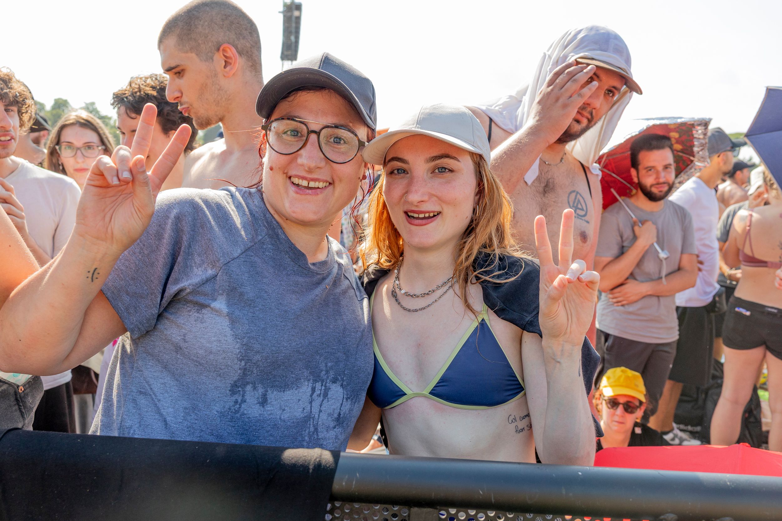 Two smiling women in caps pose for a selfie at a sunny outdoor festival with a crowded group behind them, flashing peace signs.
