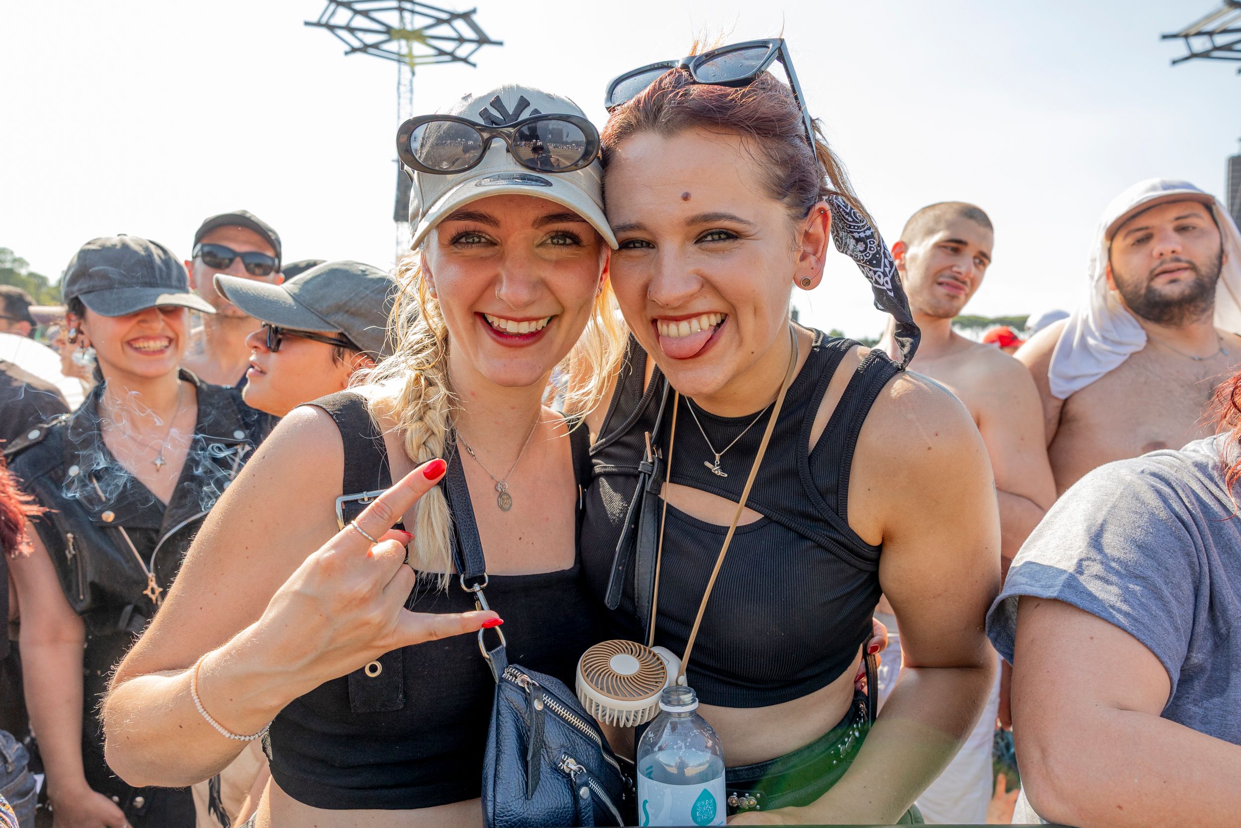 Two smiling women posing for a selfie at an outdoor festival, with sunglasses on their heads and a crowd in the background