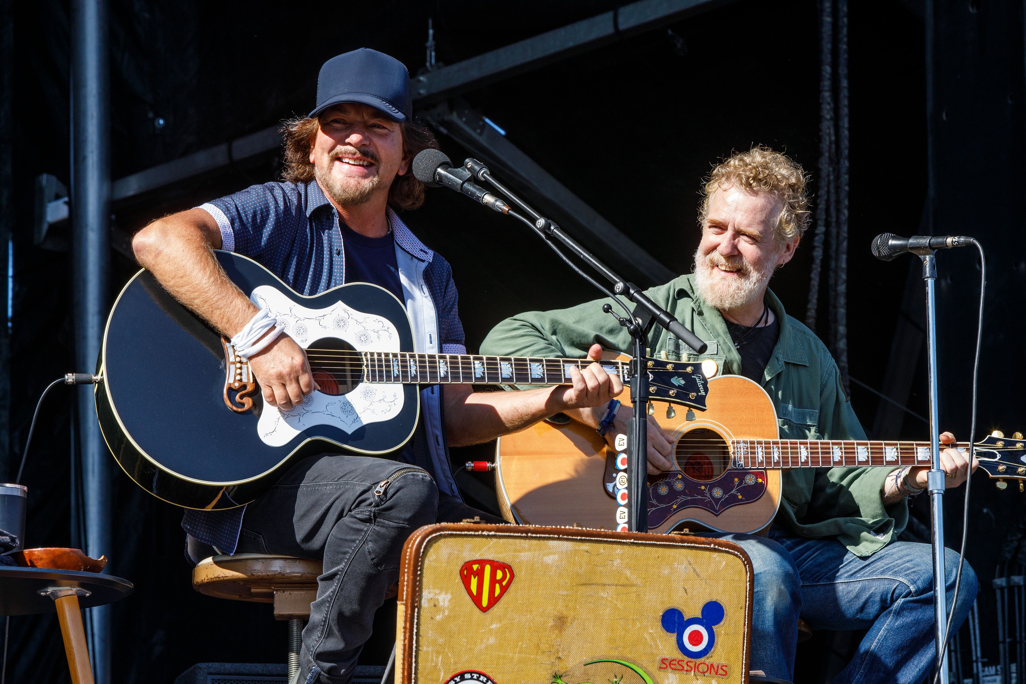 Two male musicians play acoustic guitars on a stage, singing into microphones during a live performance, smiling at the audience.