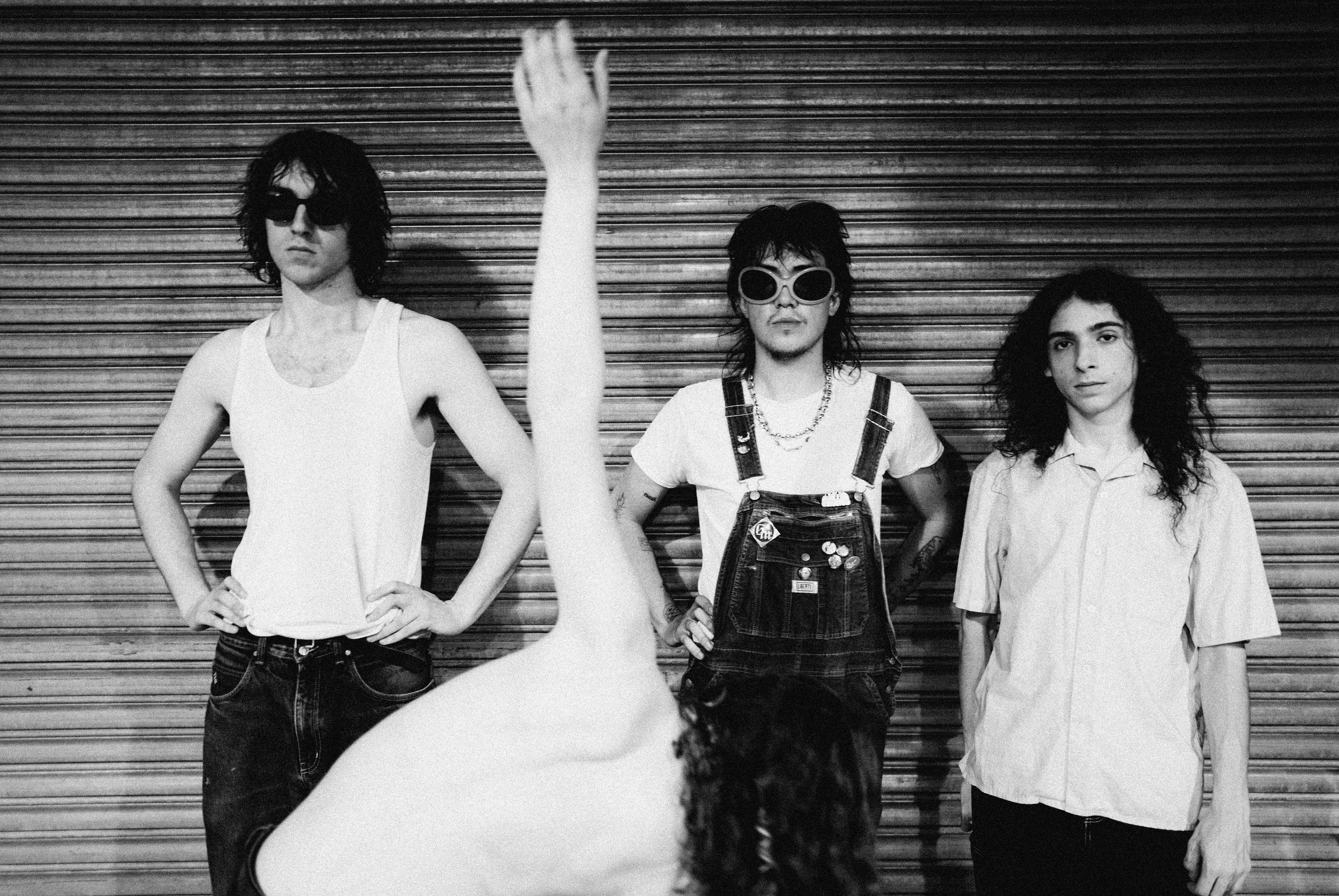 Three men pose against a metal shutter; left wears sunglasses and a white tank top, middle wears overalls and round sunglasses, right in a light shirt. (Black-and-white band photo)